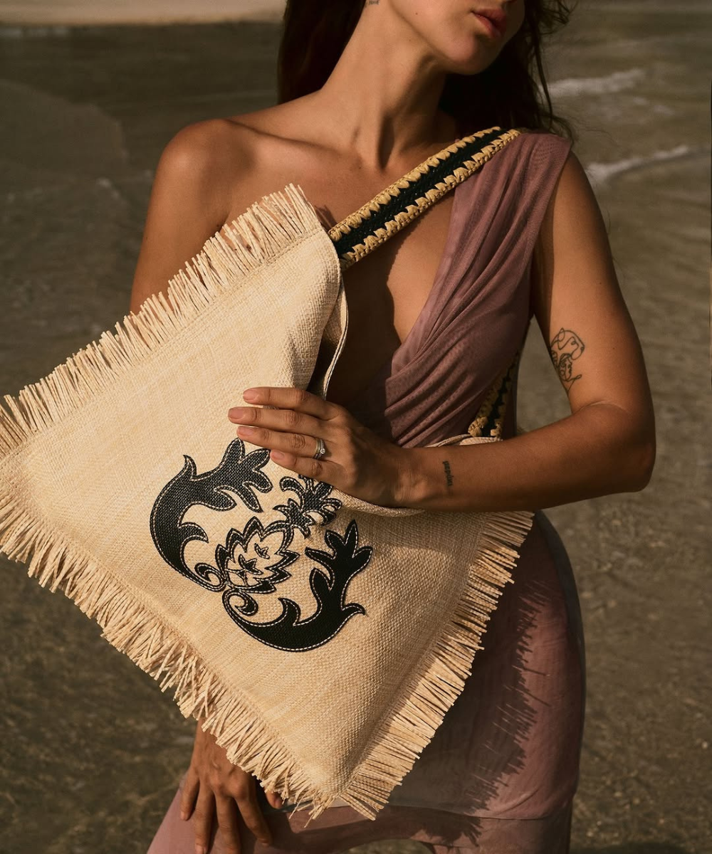 Pessoa segura bolsa de palha com estampa preta e franjas, vestindo roupa lilás em cenário de praia.