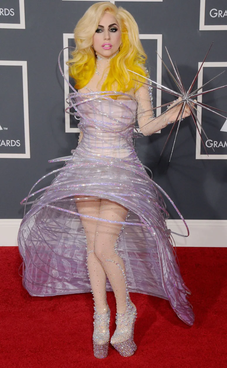 Uma pessoa com vestido futurista prateado e cabelos loiros em um evento de tapete vermelho.
