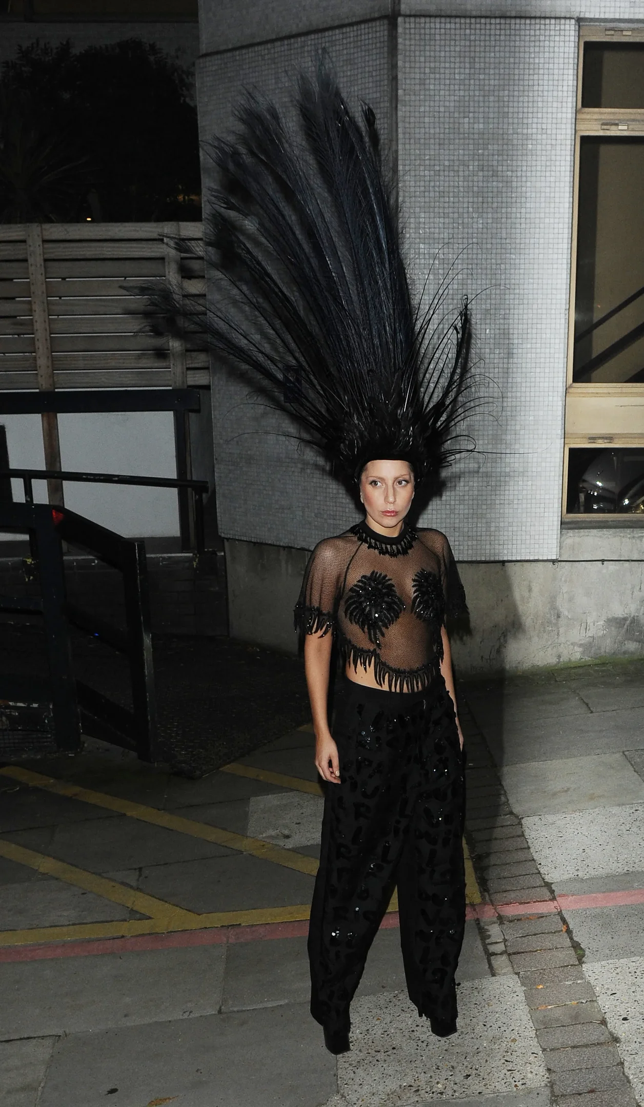 Lady Gaga usa um chapéu com penas pretas e roupa preta com detalhes brilhantes.