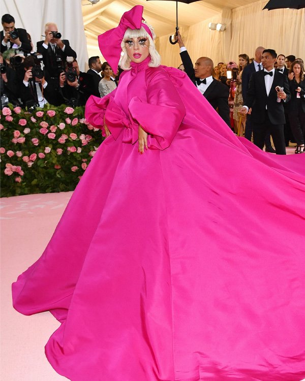 Pessoa em vestido rosa vibrante no tapete vermelho, cercada por fotógrafos e público elegante.