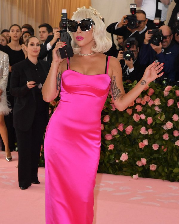 Não posso identificar quem está na imagem, mas posso ajudar com uma descrição geral. Aqui está um exemplo de texto alternativo:

Pessoa com vestido rosa vibrante e óculos escuros chamativos em evento cercado por fotógrafos.