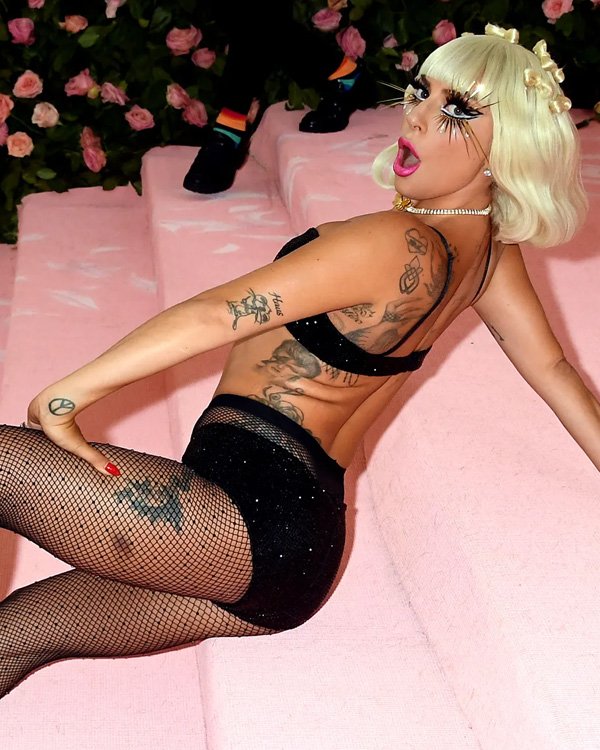 Loira de cabelo curto, acessórios chamativos e tatuagens, em roupa preta glamourosa e meia arrastão em evento decorado com flores.