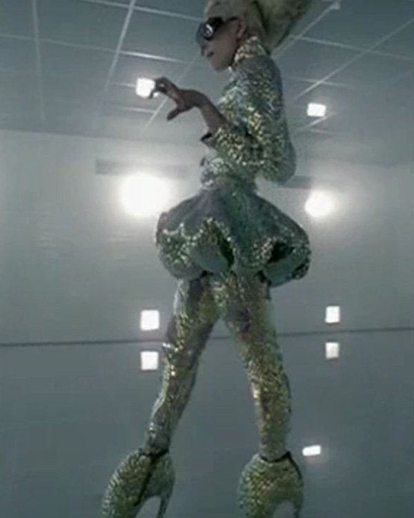 Pessoa com roupa prateada e plataformas altas num ambiente futurista, lembrando estilo Lady Gaga.