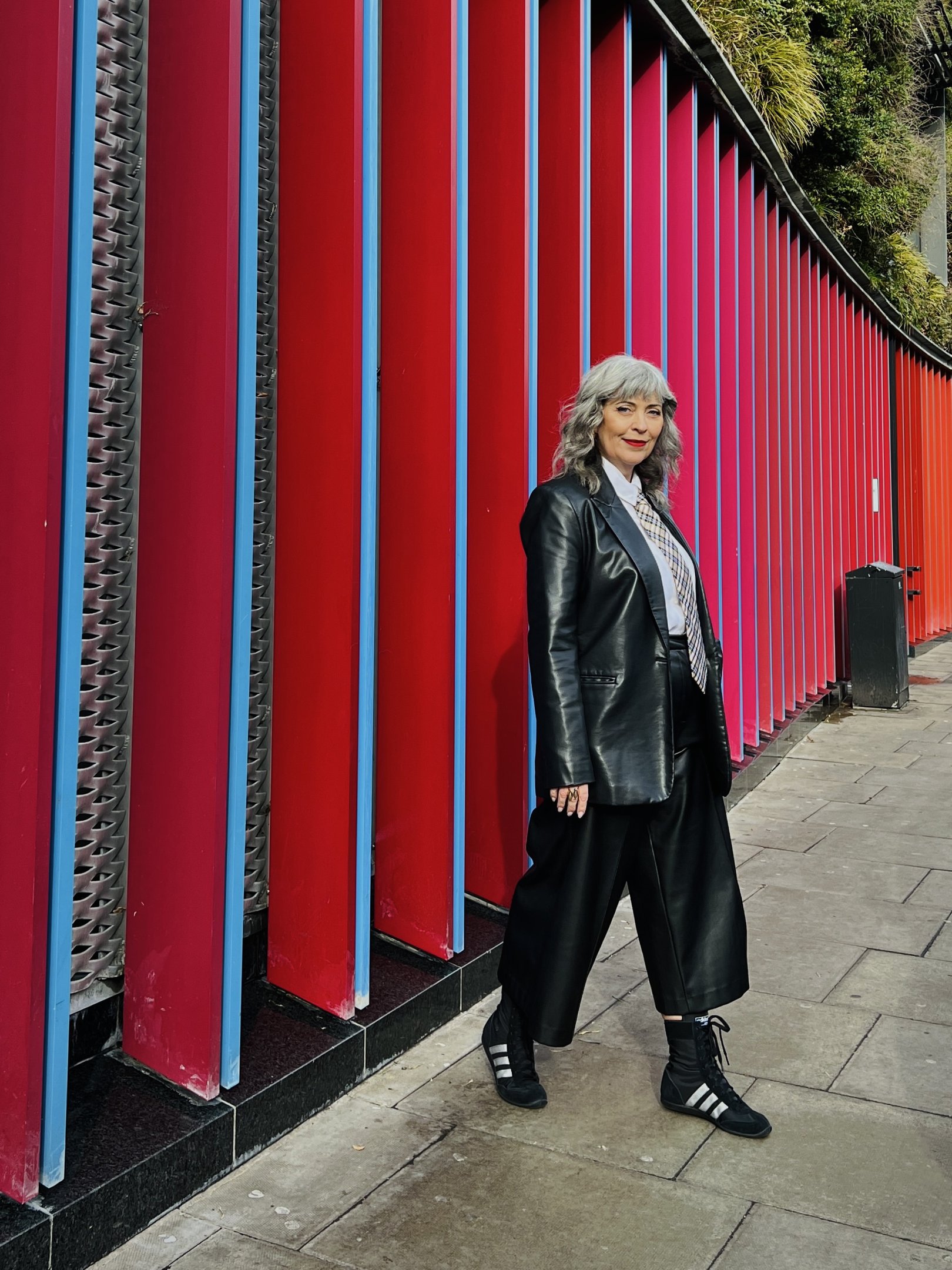 Pessoa com cabelo grisalho veste blazer preto e calças largas, caminhando ao lado de parede vermelha e azul.