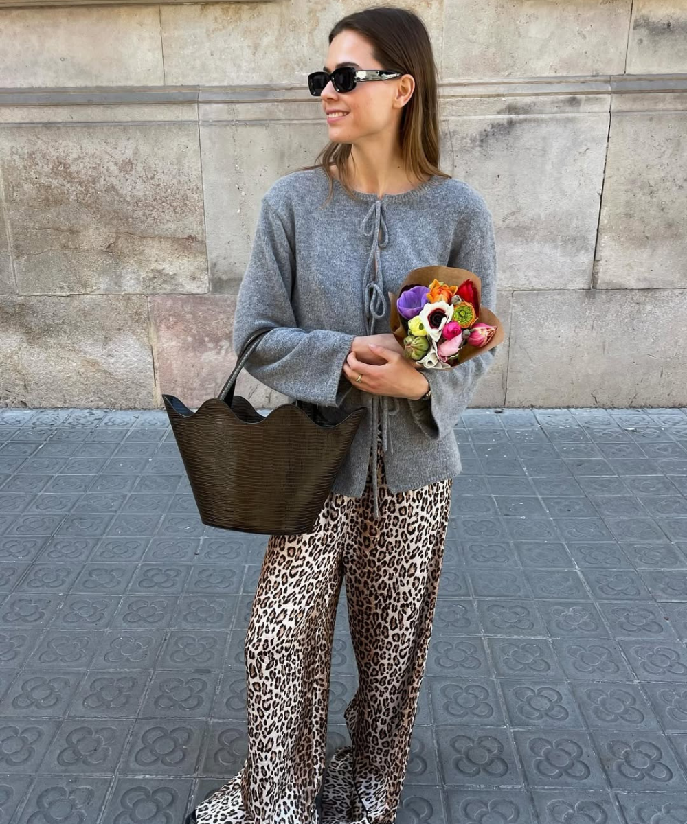 Pessoa usando calça pantalona com estampa animal e suéter cinza. Carrega bolsa preta e flores.