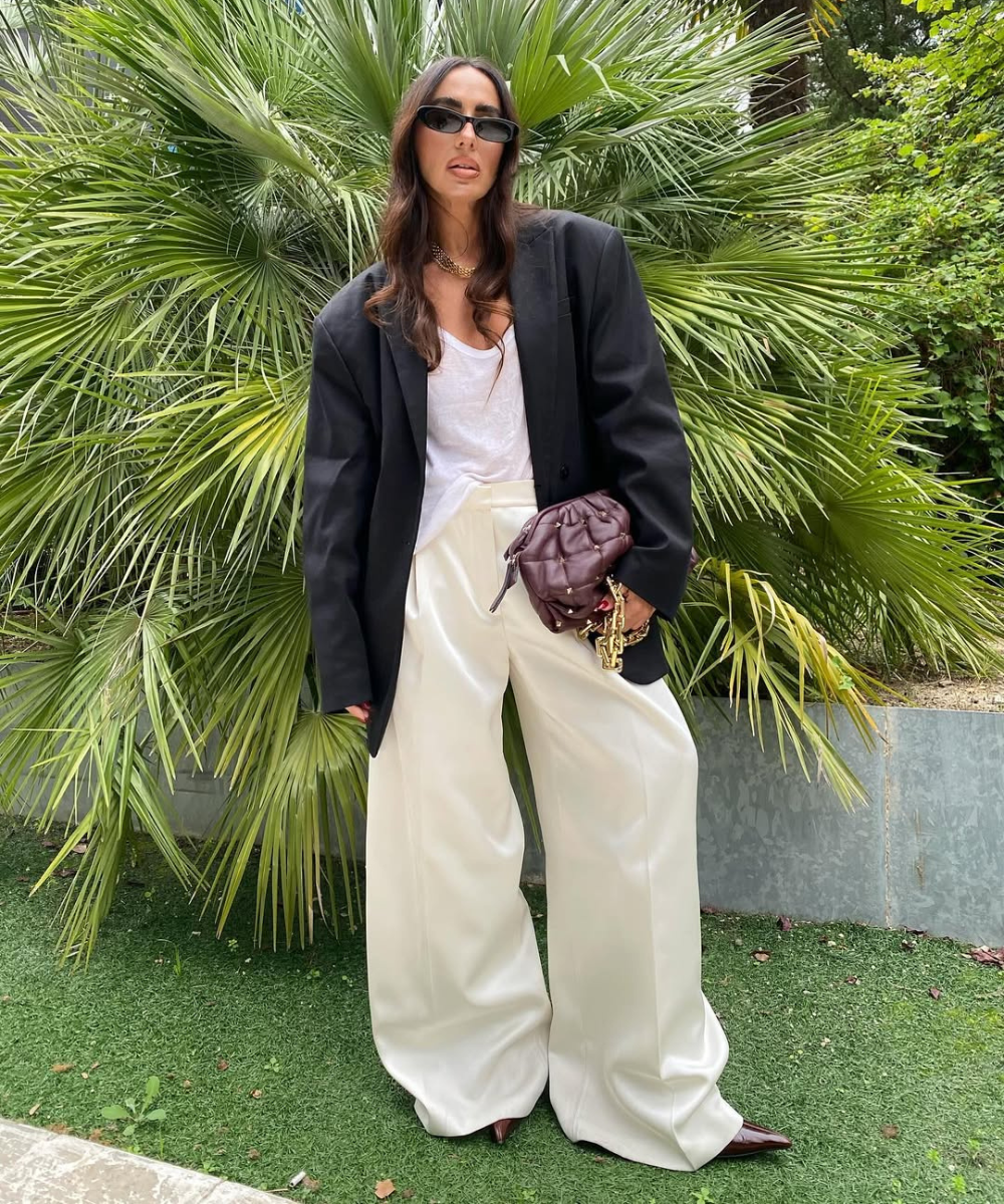 Pessoa com blazer preto e calça pantalona clara, segurando bolsa marrom, cercada por plantas verdes. Inspirações de looks com calça pantalona.