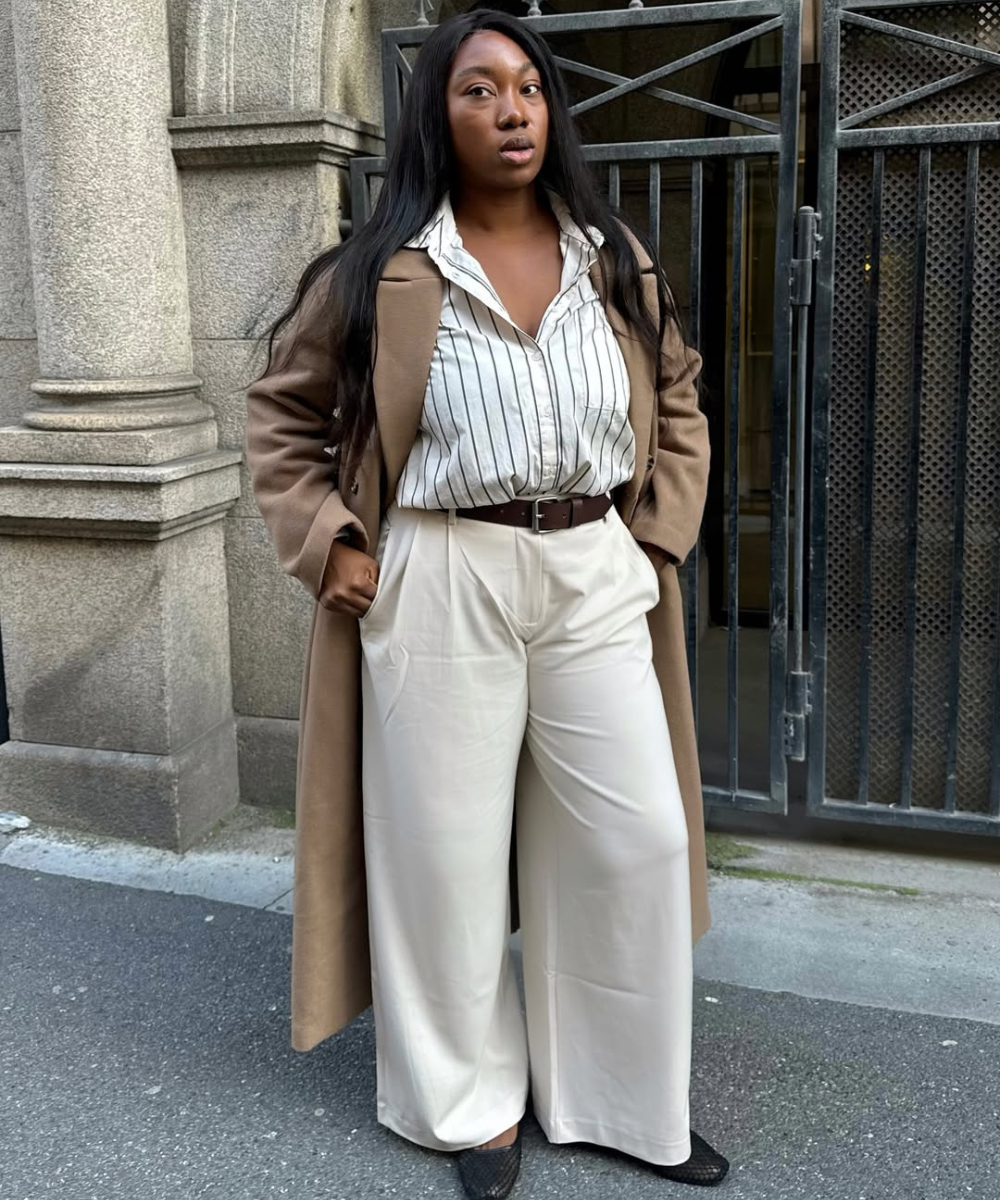 Mulher com calça pantalona branca, camisa listrada e casaco marrom. Looks elegante em cenário urbano.