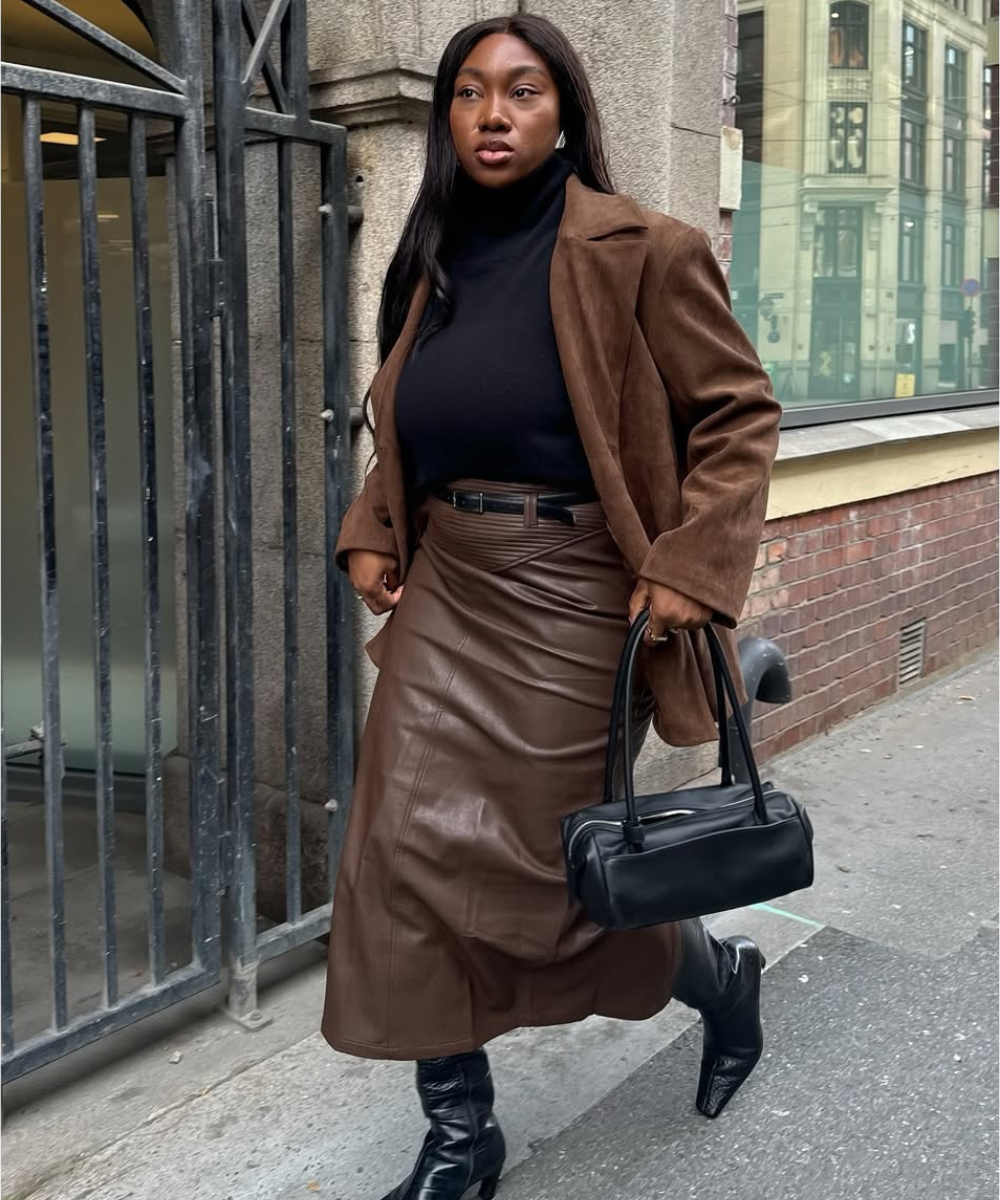 Pessoa caminhando, usando casaco e saia de couro marrom, blusa preta e botas pretas, segurando bolsa preta.