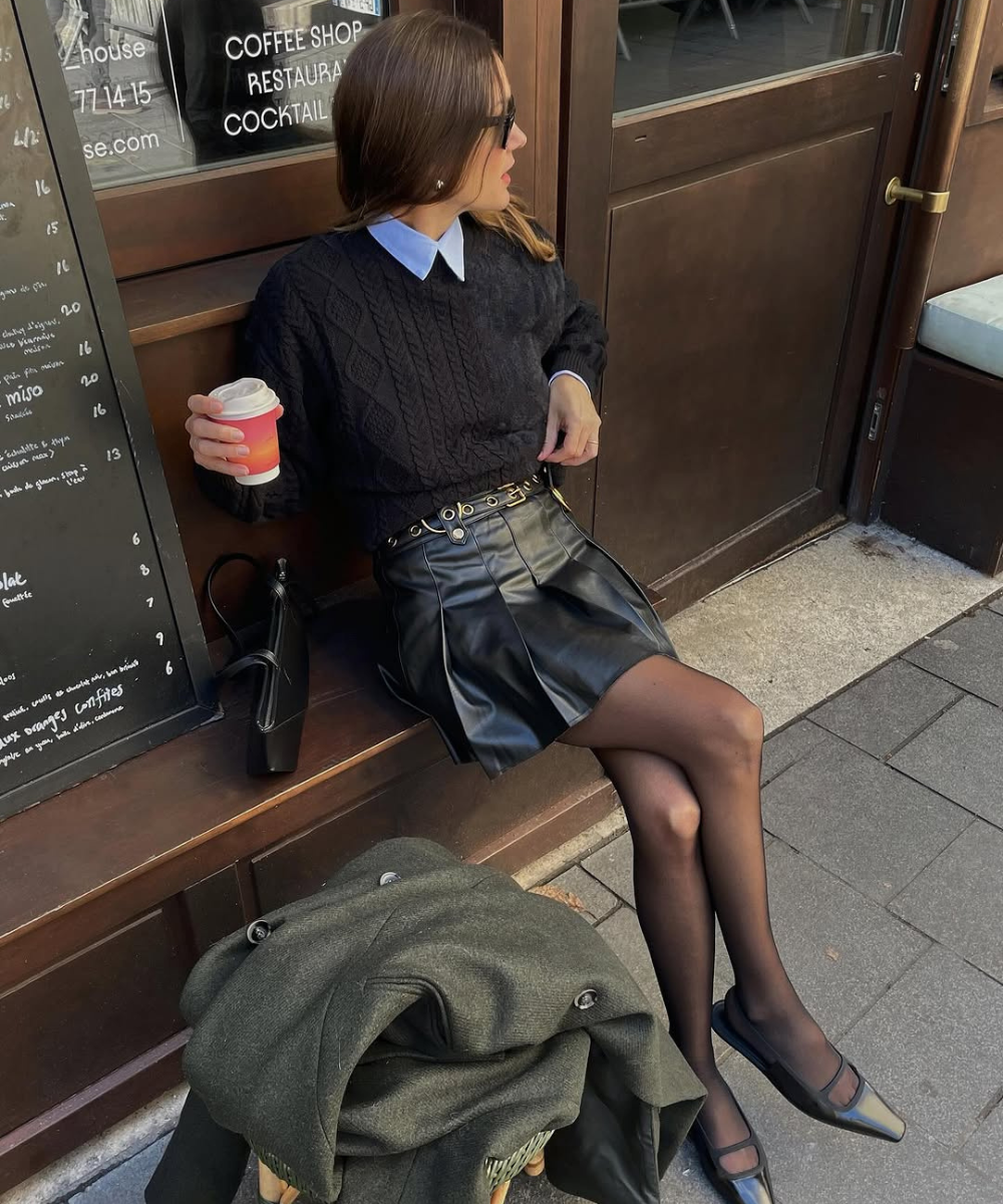 Mulher sentada em banco, vestindo saia de couro preta e suéter escuro, segurando café.