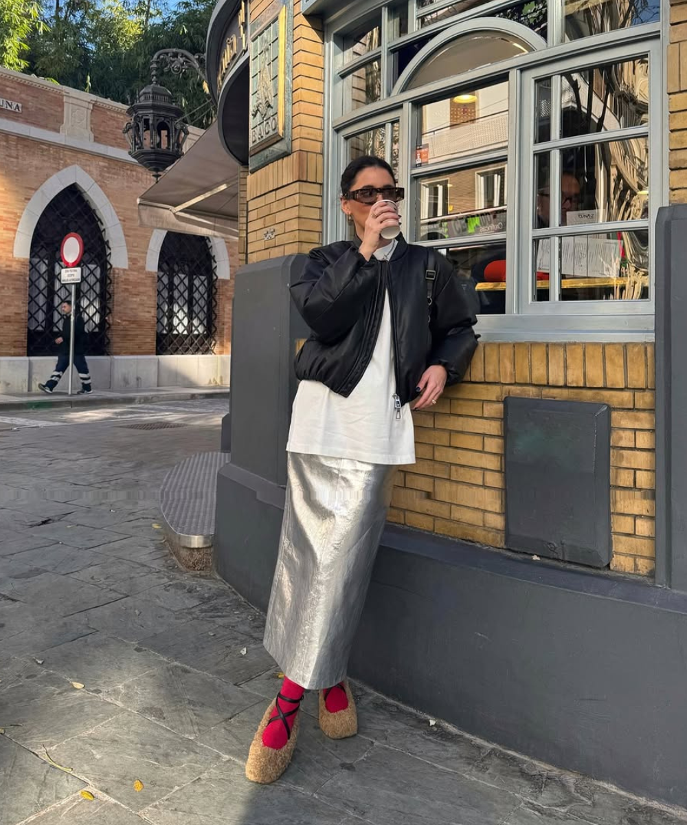 Pessoa vestindo jaqueta preta e saia metalizada, tomando café em calçada urbana. Inspirações de looks com saia de couro.