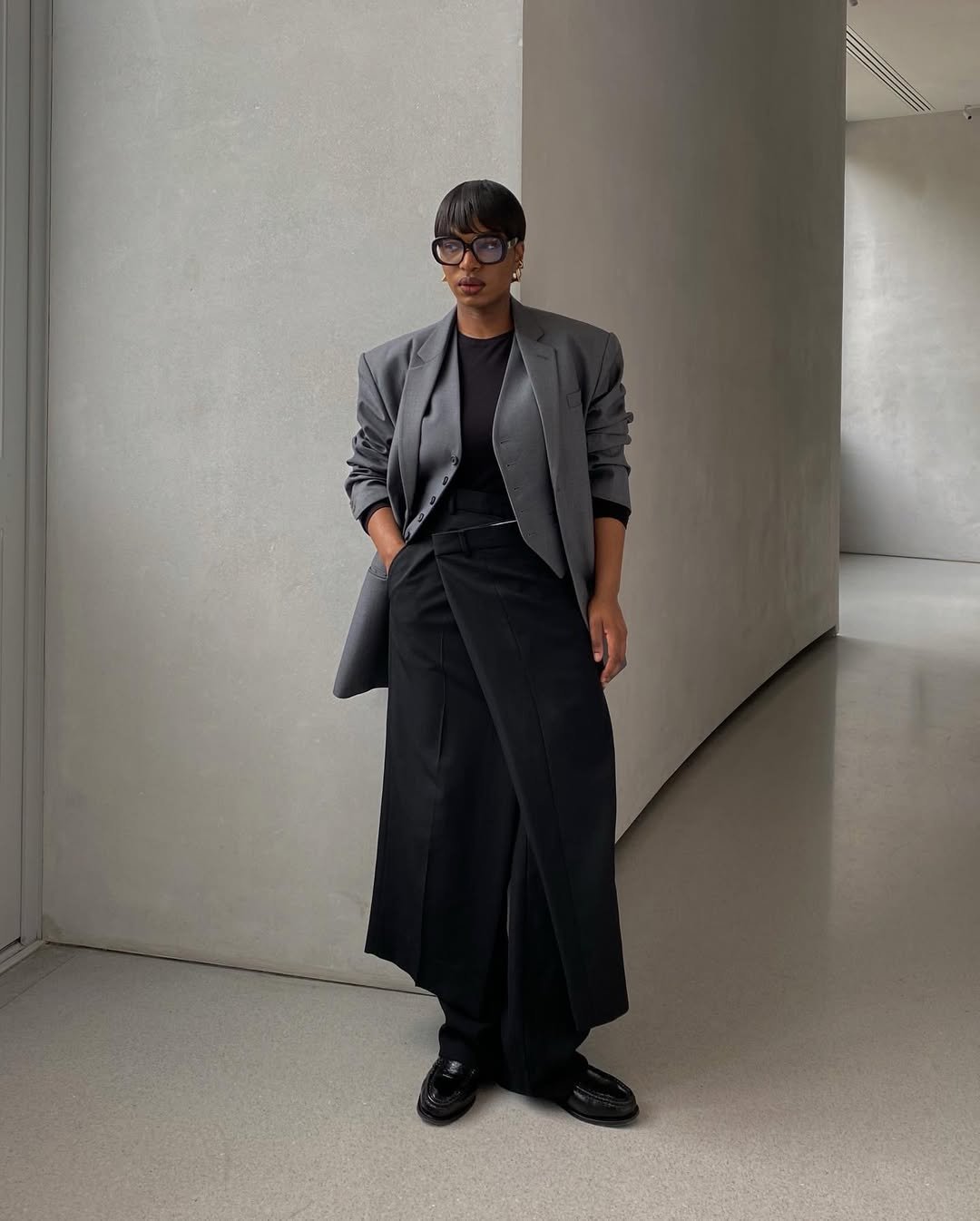 Pessoa de maxi blazer cinza, blusa preta e calça larga, em pé em um corredor minimalista.