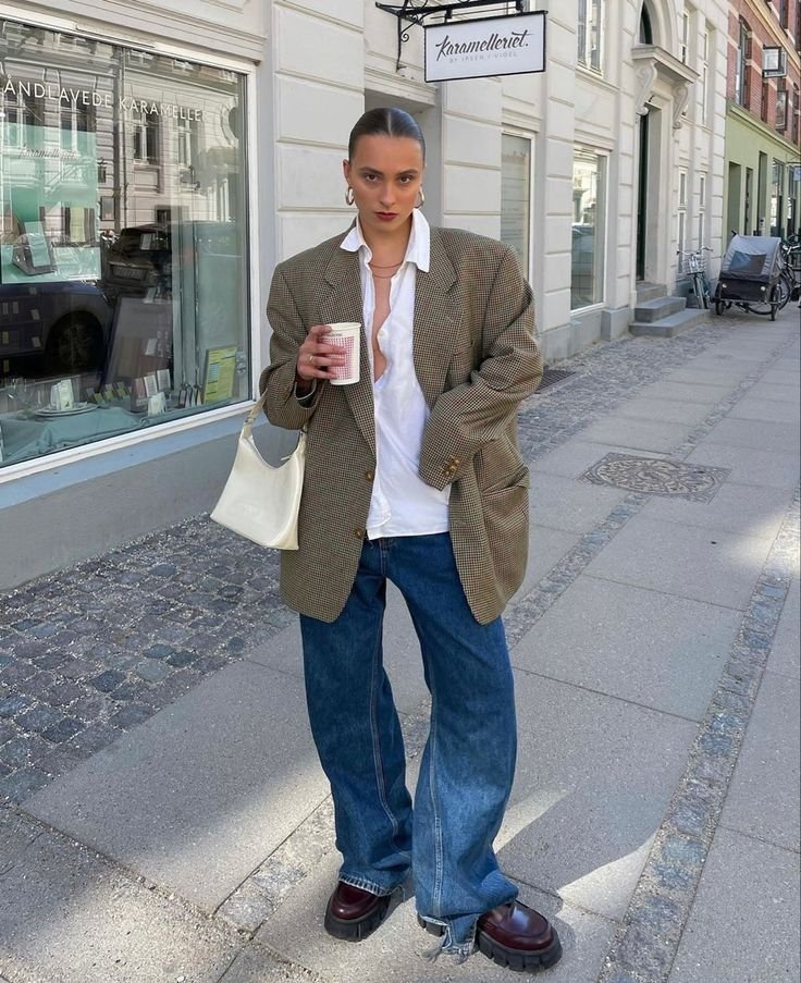 Pessoa usando maxi blazer xadrez, camisa branca e calças jeans largas em rua urbana. Segura bebida e bolsa clara.