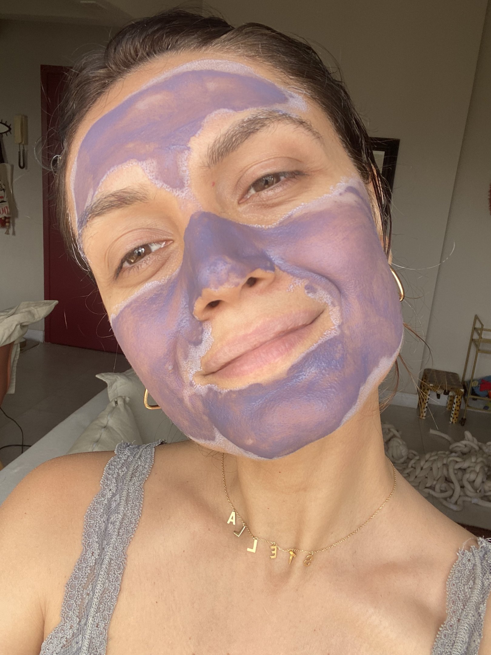 Pessoa sorri enquanto usa máscara de argila da benefit facial roxa, no ambiente interno, vestindo colar dourado e blusa cinza.