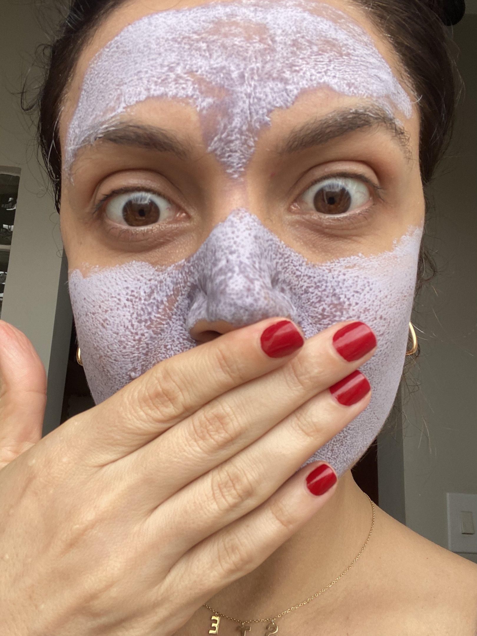 Mulher com máscara de argila da benefit  roxa e unhas vermelhas cobrindo a boca.