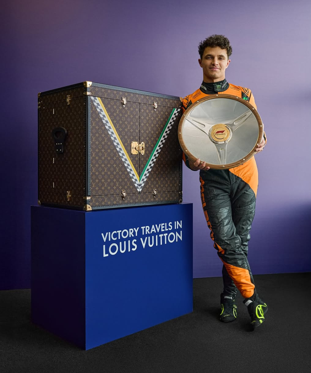 Piloto de Fórmula 1 em macacão laranja com troféu e baú Louis Vuitton, destacando a moda na categoria.