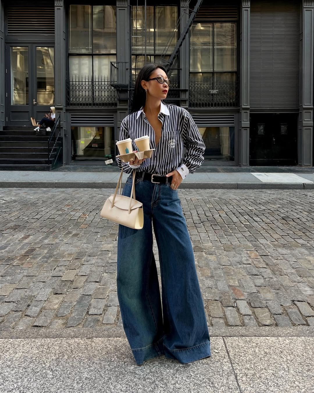 Pessoa com camisa listrada e calça jeans larga tendência, segura bebidas e bolsa em rua de paralelepípedos.