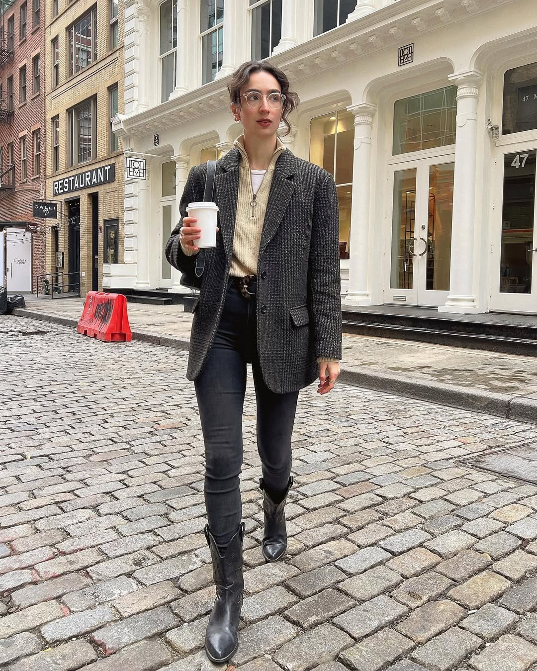 Pessoa caminhando em rua urbana usando calça jeans preta, casaco comprido e botas, segurando café. Moda outono casual.