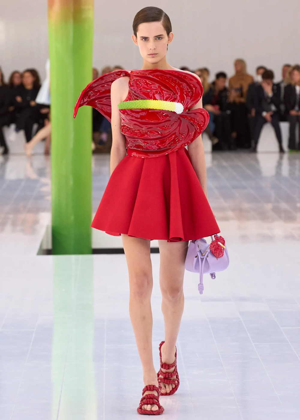 Desfile de moda com modelo usando vestido vermelho com detalhe escultural, segurando uma bolsa lilás.