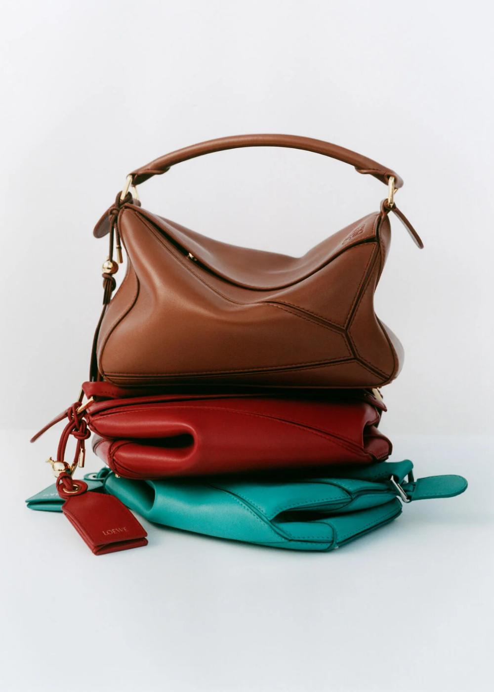 Bolsas empilhadas em marrom, vermelho e azul-turquesa, possivelmente associadas ao design de Jonathan Anderson.