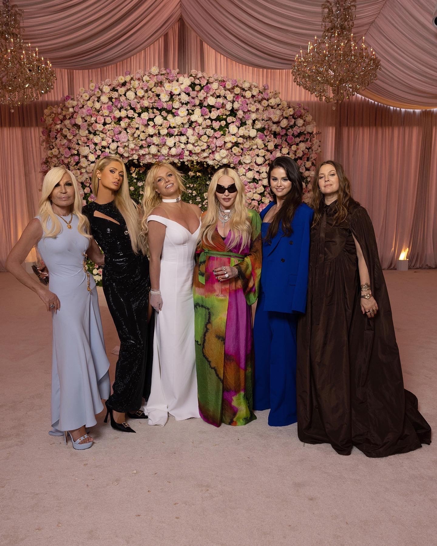 Seis mulheres posam sorridentes em um evento elegante, cercado por decoração floral luxuosa e iluminação suave.