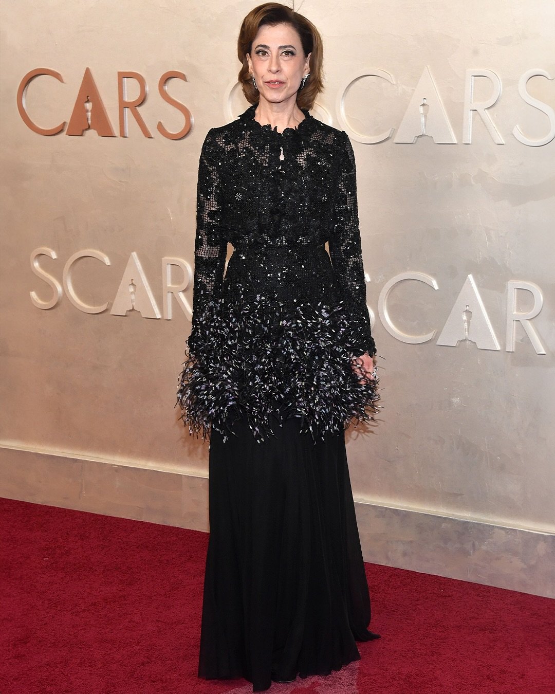 Pessoa em traje de gala preto no tapete vermelho do Oscar 2025, fundo com o logotipo do evento.