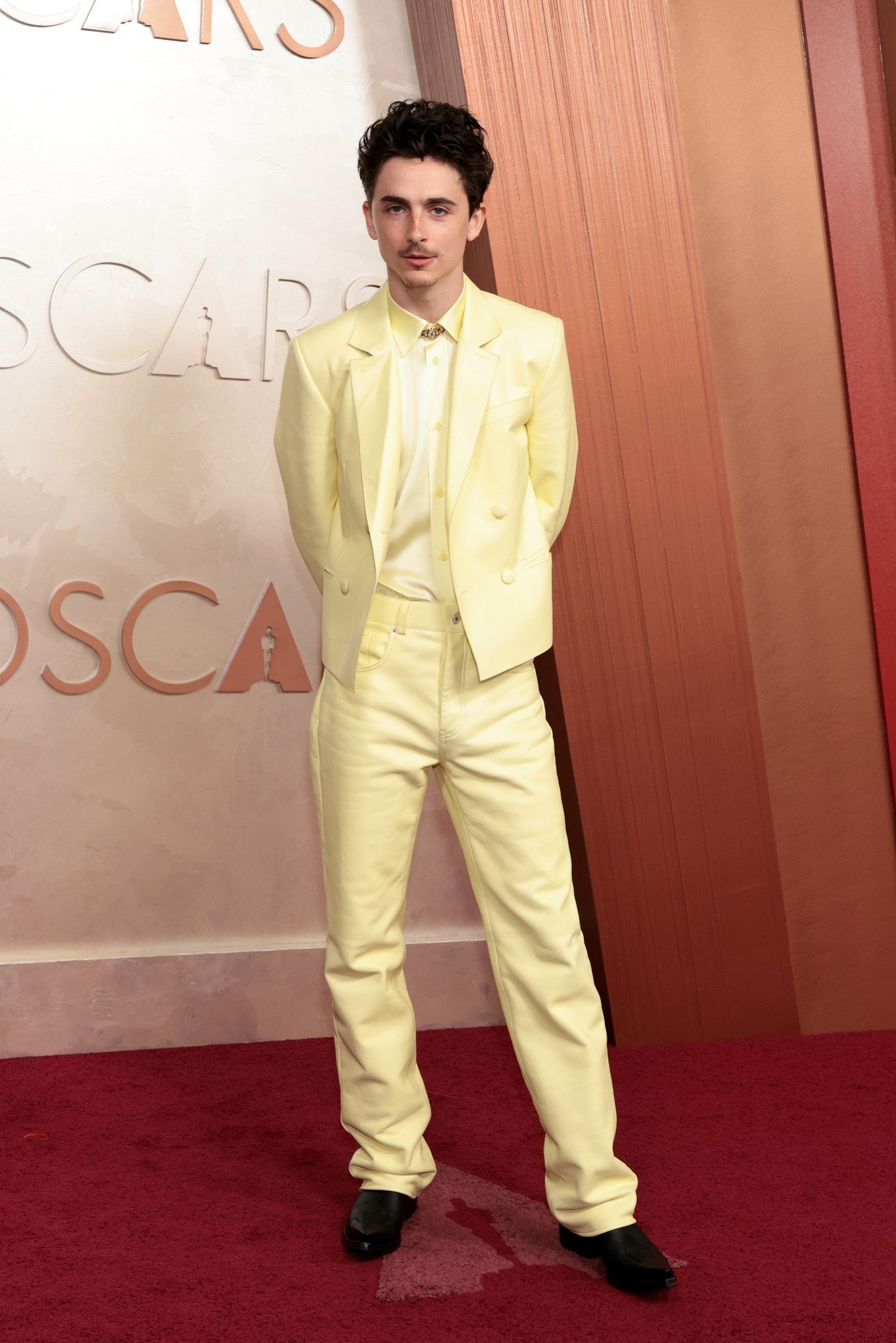 Homem em traje amarelo pálido em tapete vermelho, com logotipo do Oscar 2025 ao fundo.
