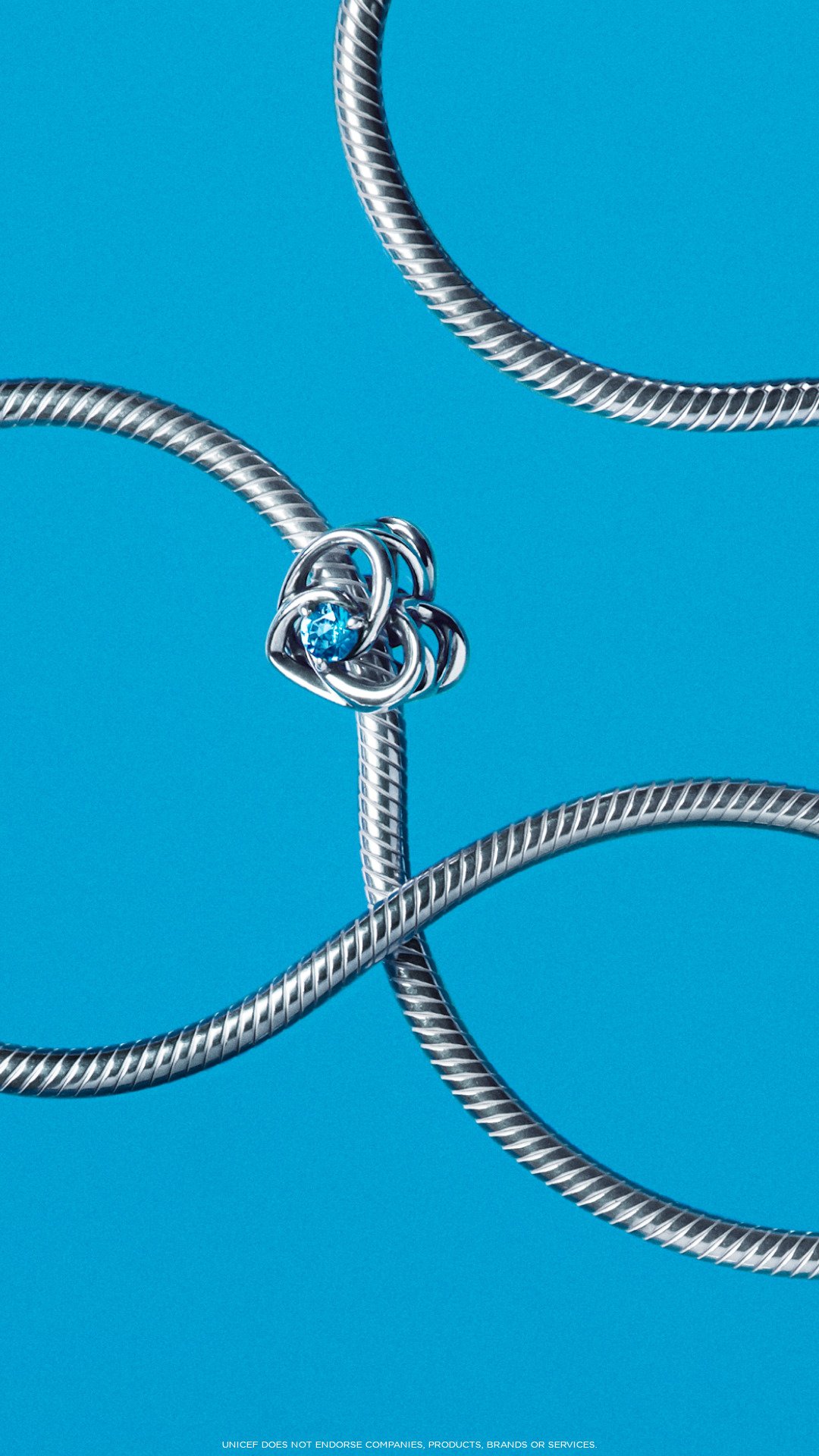 Pulseira prata entrelaçada com um ornamento floral e pedra azul no centro. Fundo azul vibrante. Pandora e UNICEF.