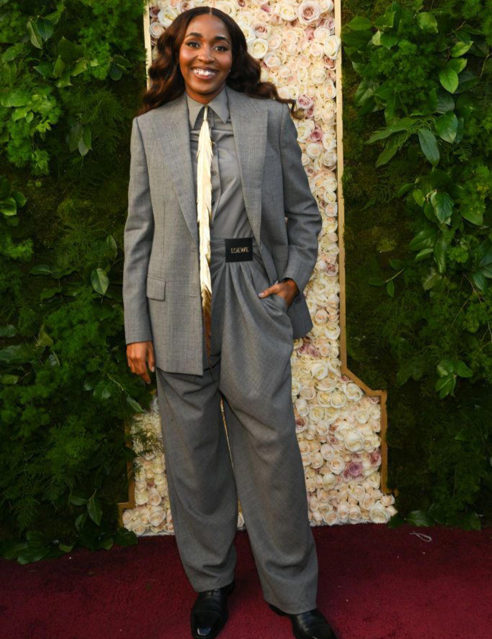Pessoa em traje cinza elegante com gravata alternativa, posando em frente a parede floral, referências na moda.
