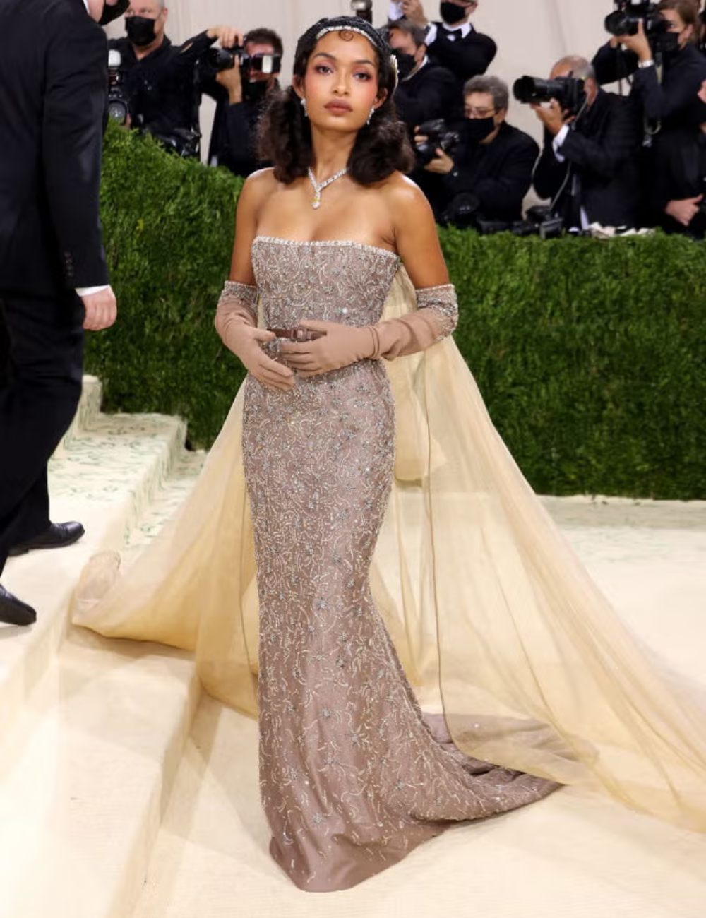 Vestido dourado elegante com detalhes em joias e capa delicada, capturando referências na moda em evento glamoroso.
