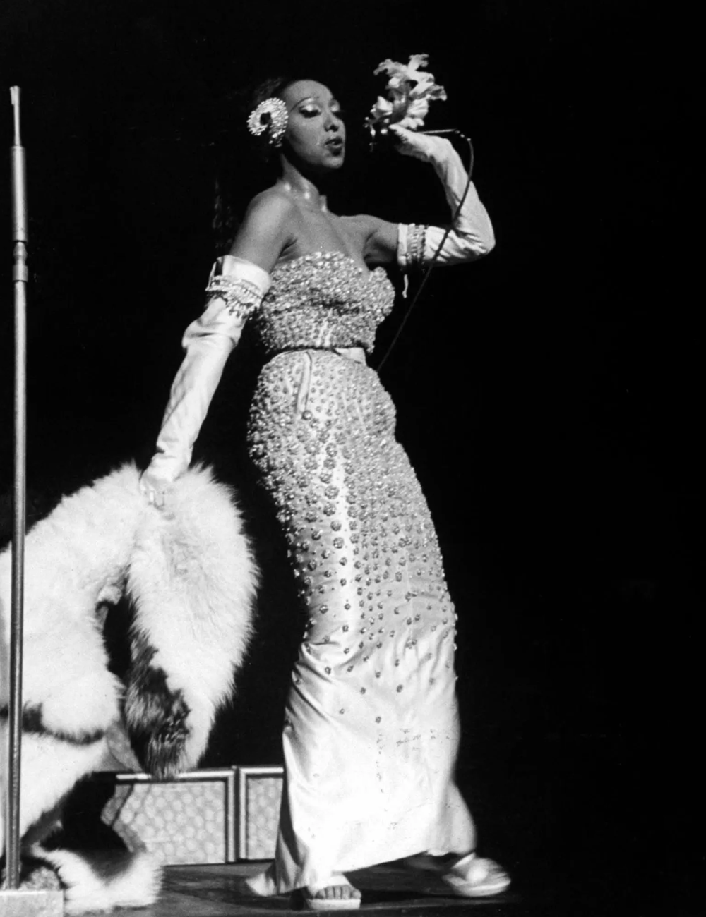 Cantora glamourosa performa em palco, usando vestido brilhante com joias e segurando estola de pele. Referências na moda.