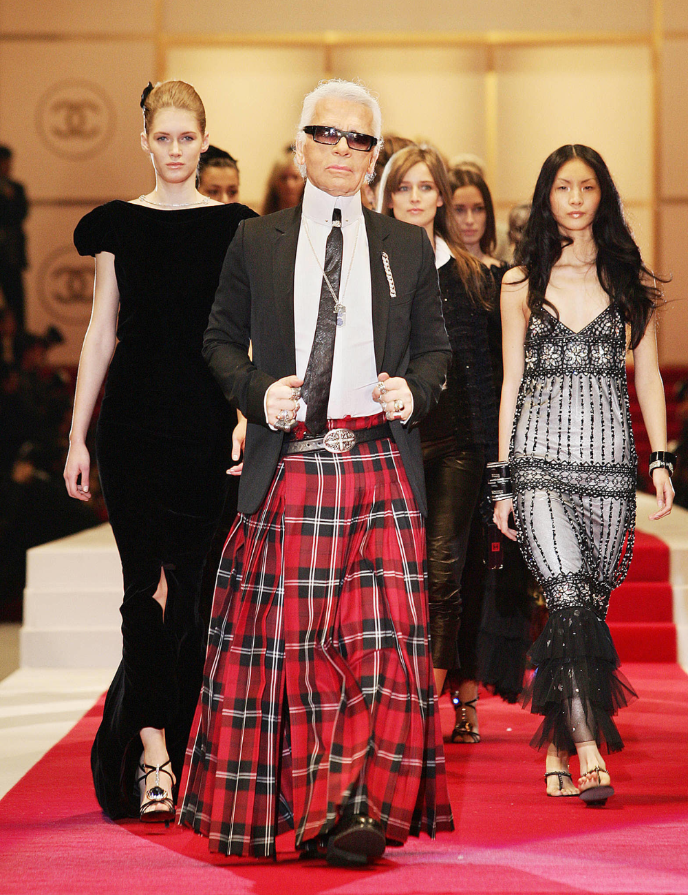 Desfile de moda com destaque em estilista com kilt xadrez e modelos em vestidos sofisticados.