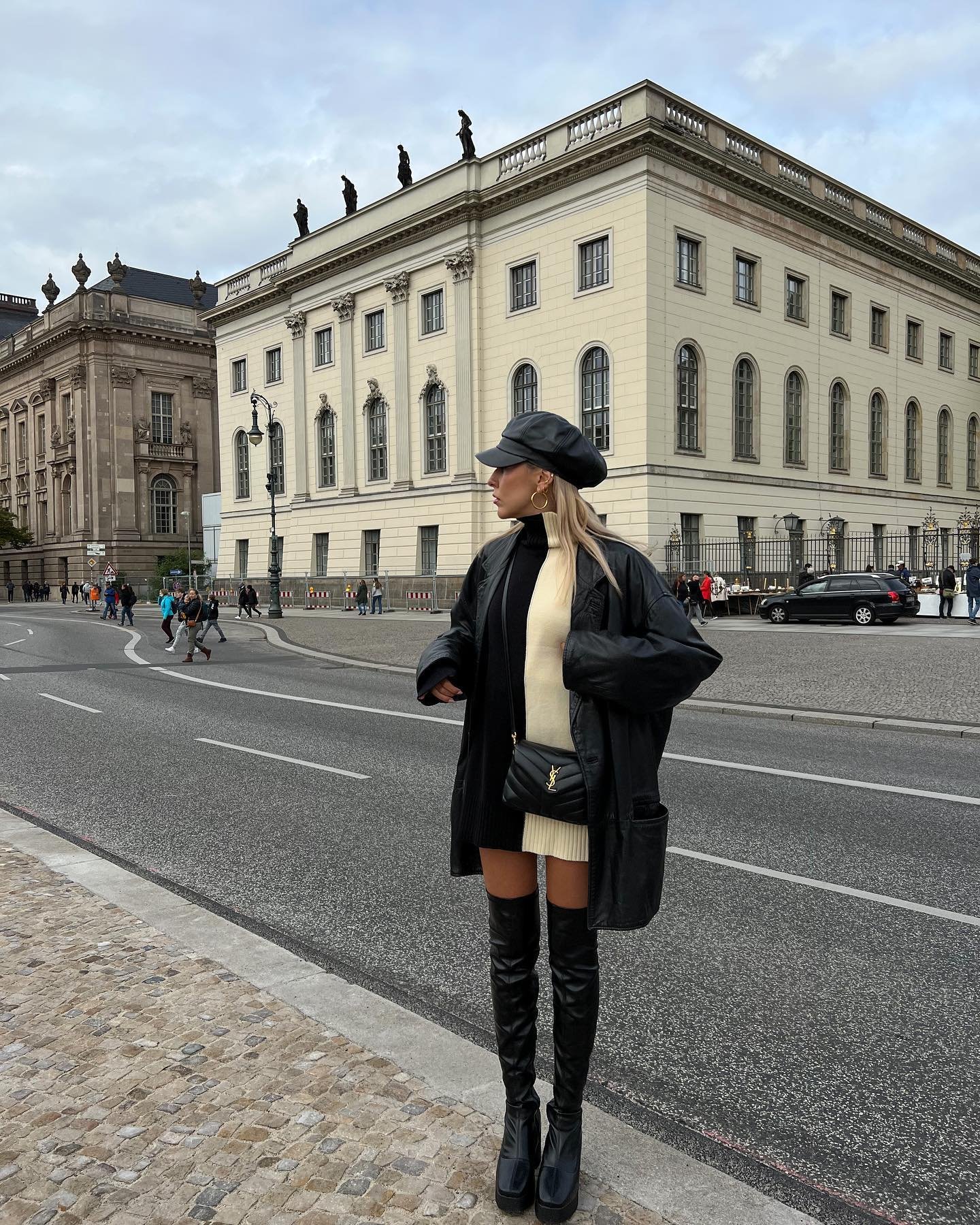 Modelo usa botas de cano alto e jaqueta preta em frente a prédio histórico. Os sapatos retrô complementam o look.