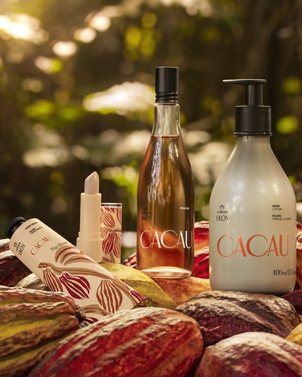 Produtos Natura Ekos Cacau: creme, batom, loção e perfume sobre cacaus em fundo de floresta iluminada.