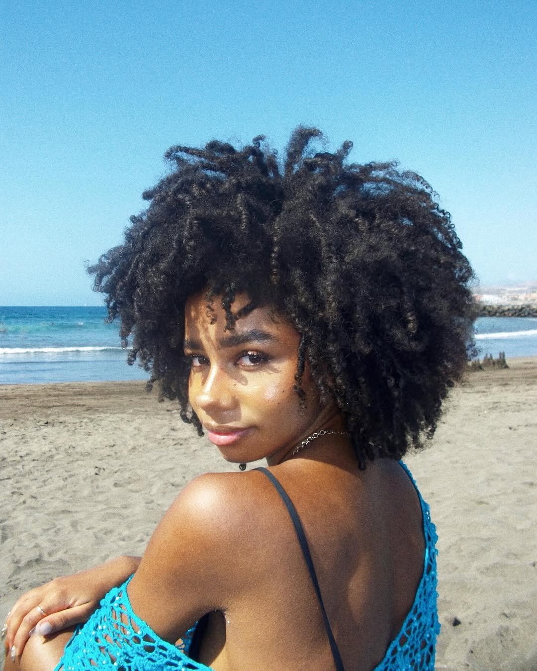 Mulher na praia com cortes de cabelo crespo e cacheado volumoso, olhando para trás e usando roupa azul.