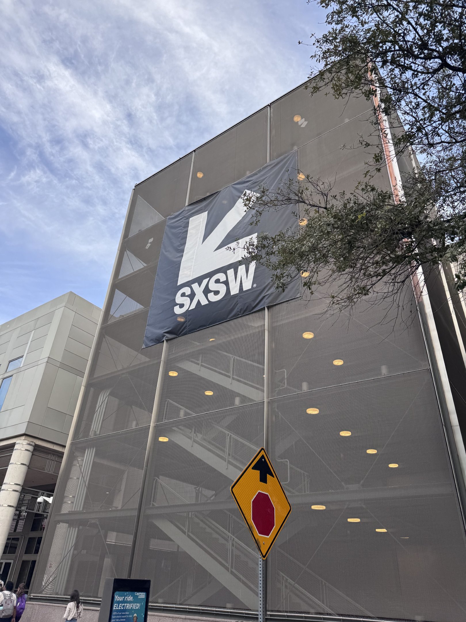 Banner grande do SXSW pendurado em edifício, mostrando escadas internas sob céu azul com árvore ao lado.