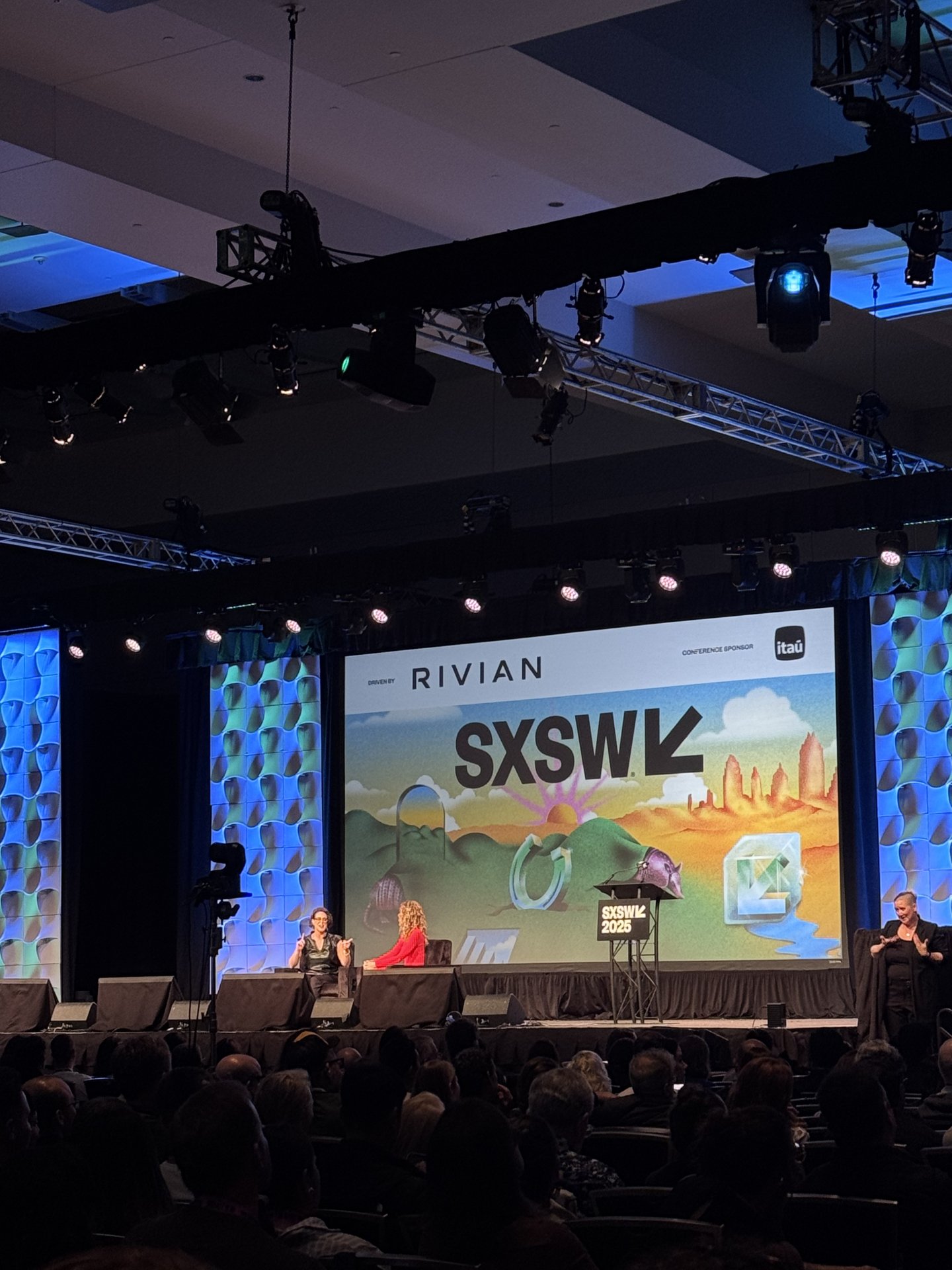 Palestra do SXSW 2025 com duas mediadoras no palco, telão ao fundo com logotipo e público assistindo.