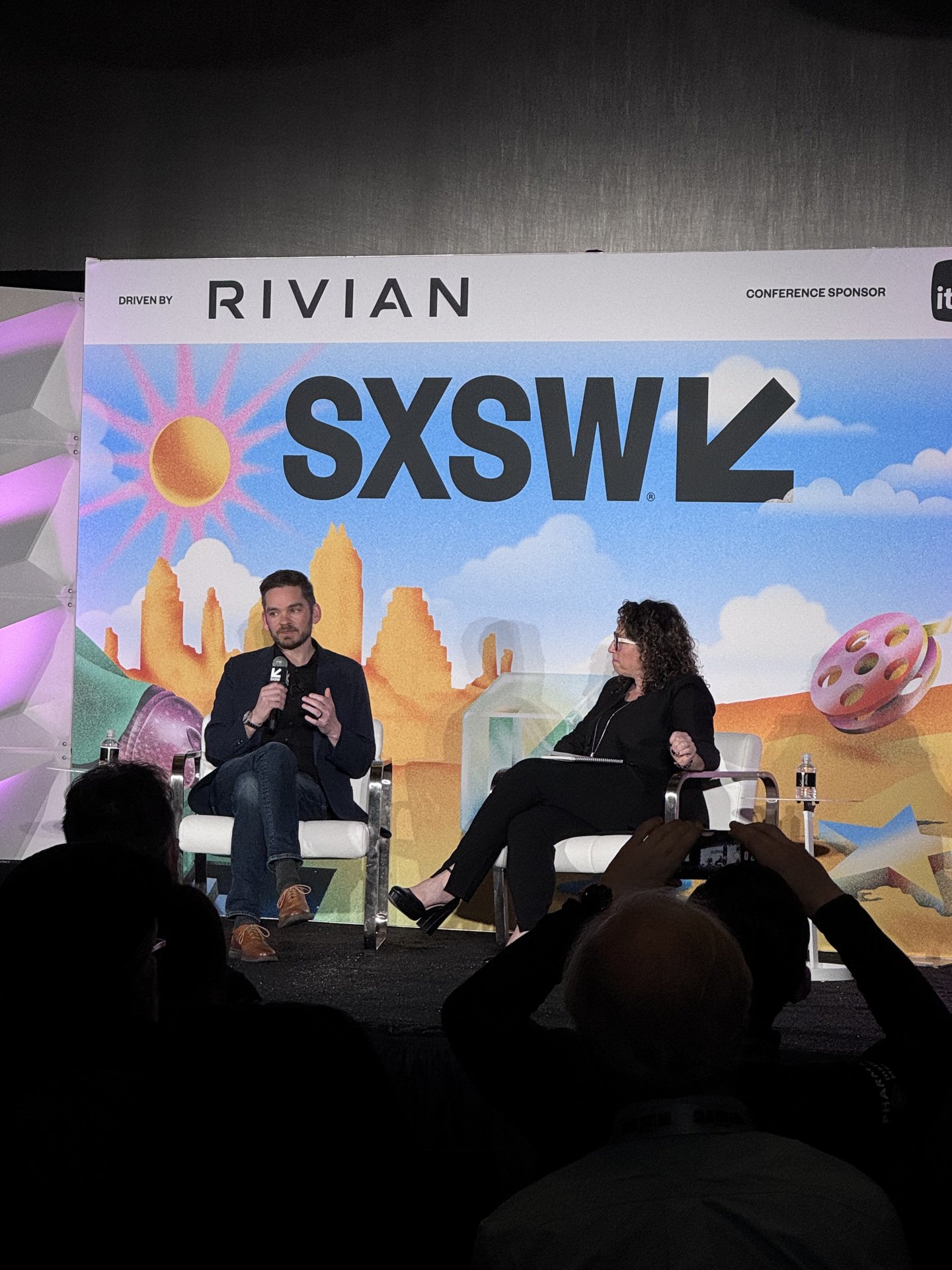 Dois palestrantes discutem em um painel sobre SXSW 2025, com banner colorido ao fundo.