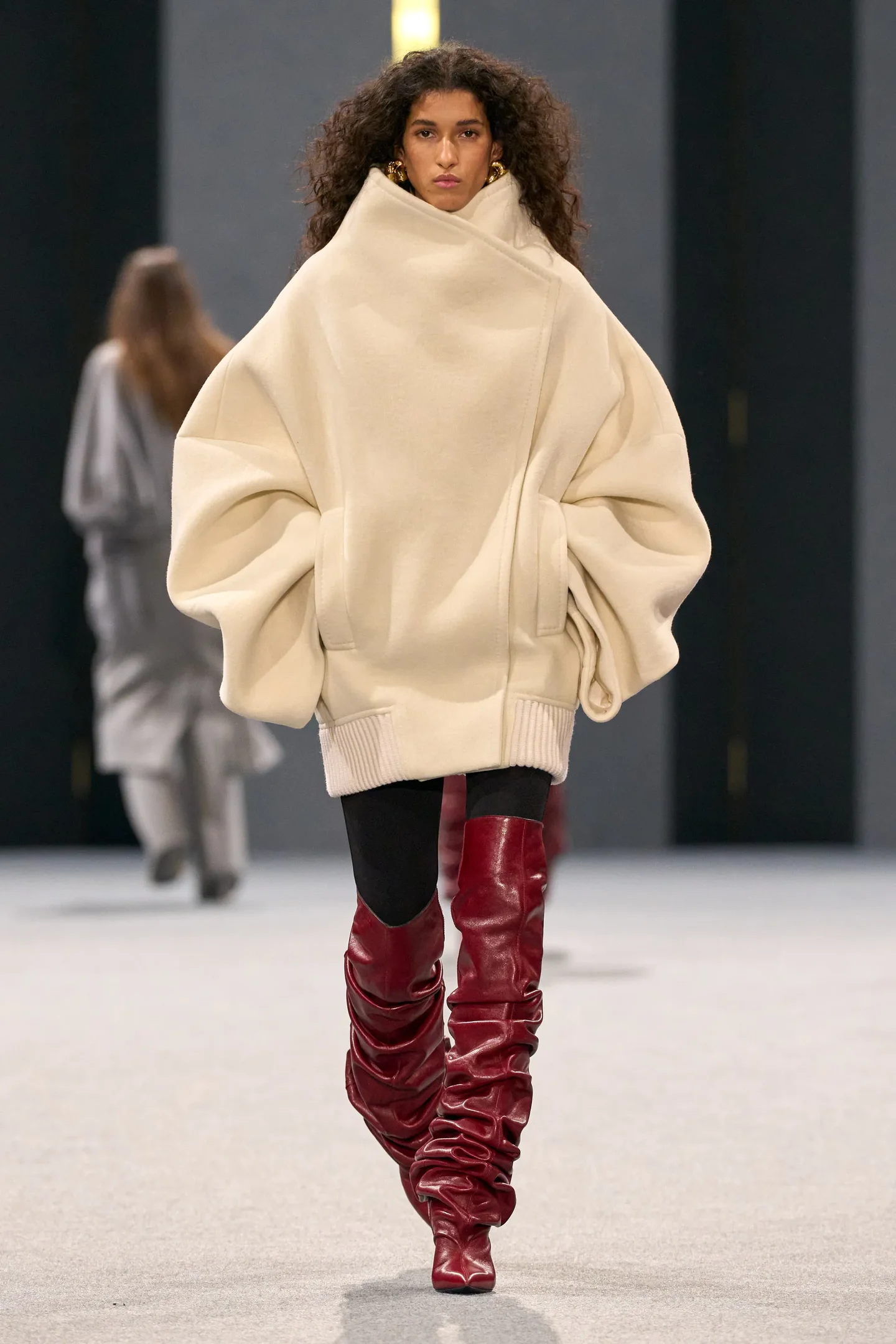 Modelo veste casaco oversized bege e botas vermelhas, destacando-se na passarela da temporada Outono/Inverno 25.