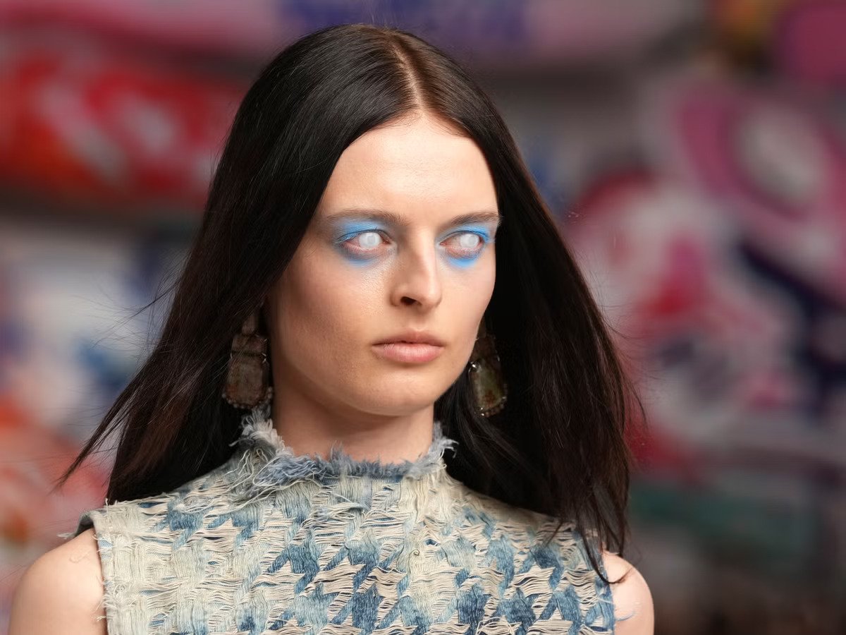 Modelo com maquiagem azul vibrante caminha em desfile, destacando tendência ousada no mês da moda.