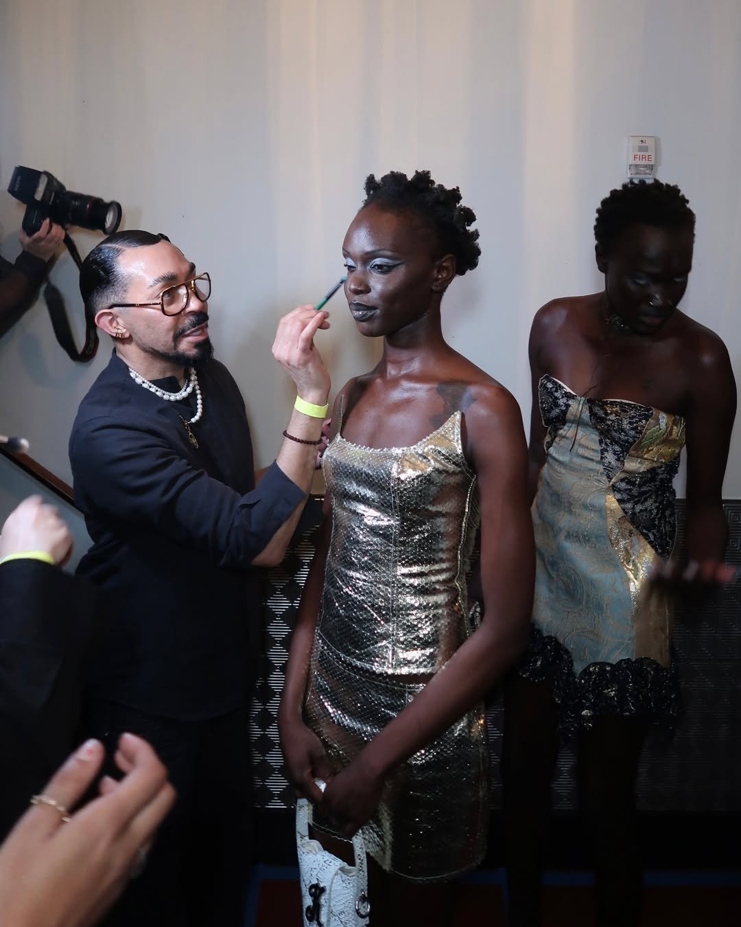 Maquiador aplica batom em modelo com vestido dourado; tendências de beleza em destaque.