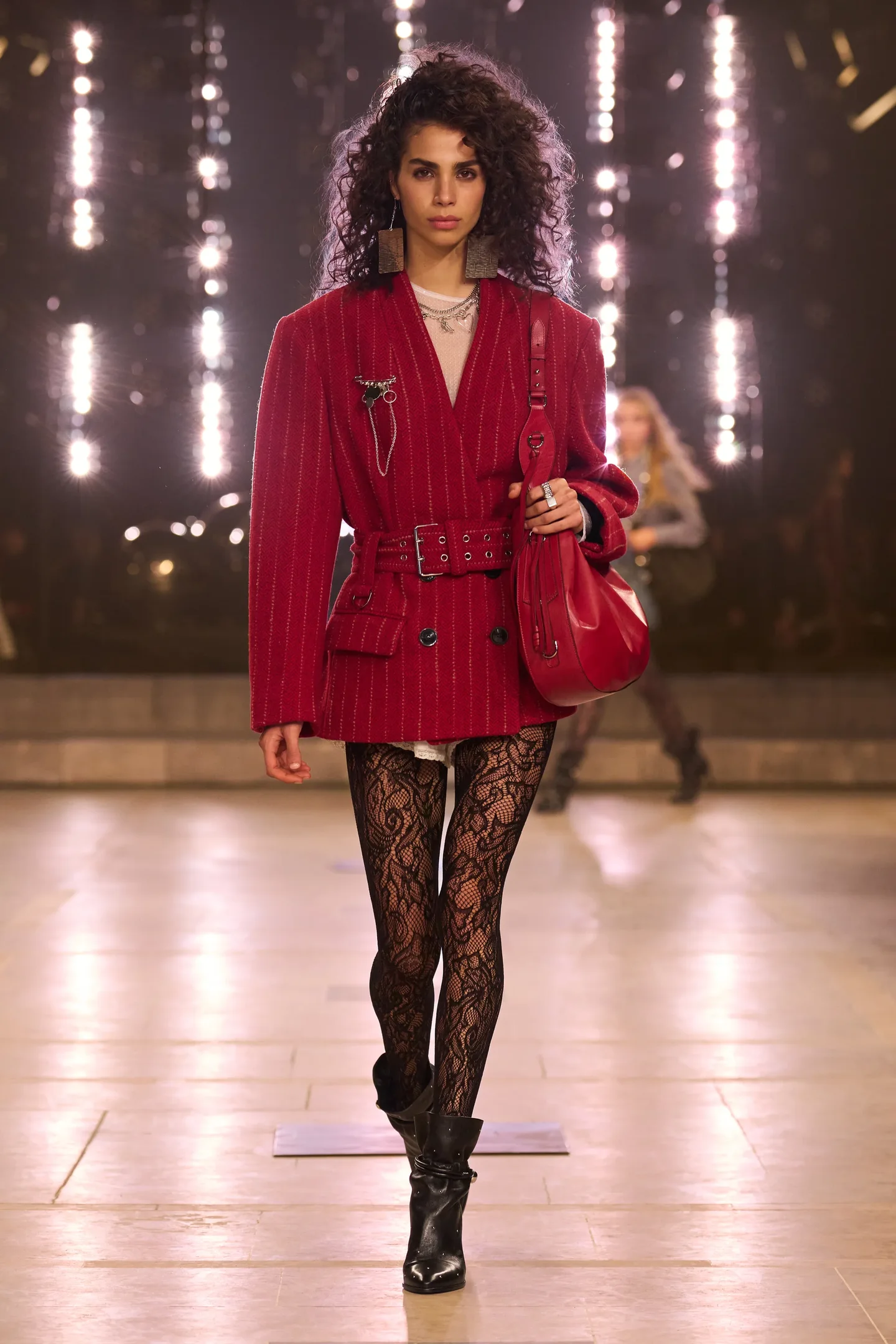 Modelo desfila com blazer vermelho, meia-calça rendada e botas pretas, refletindo tendências da temporada Outono/Inverno 25.