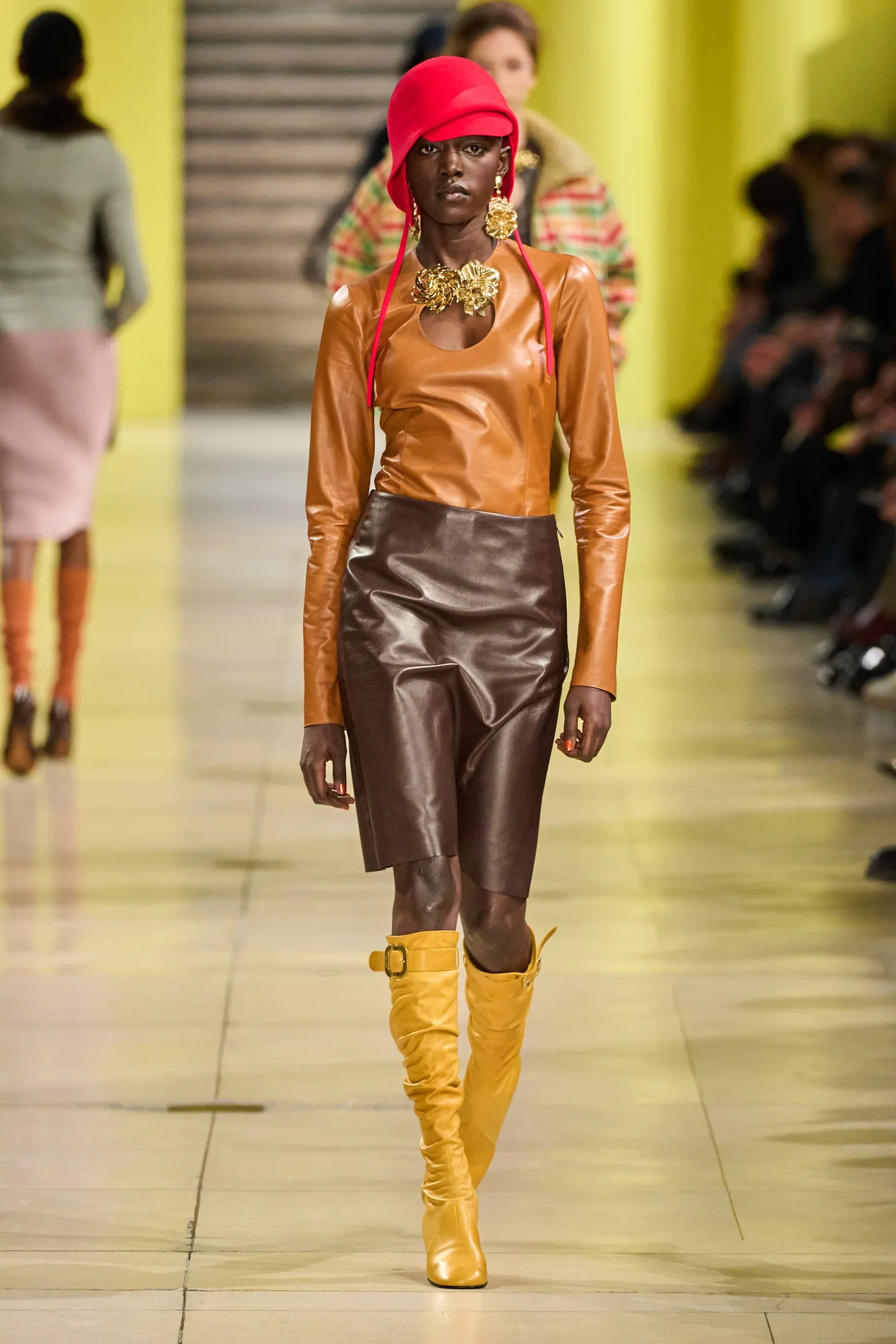 Modelo desfila com look em couro marrom e amarelo, chapéu vermelho e botas amarelas, tendência Outono/Inverno 25.