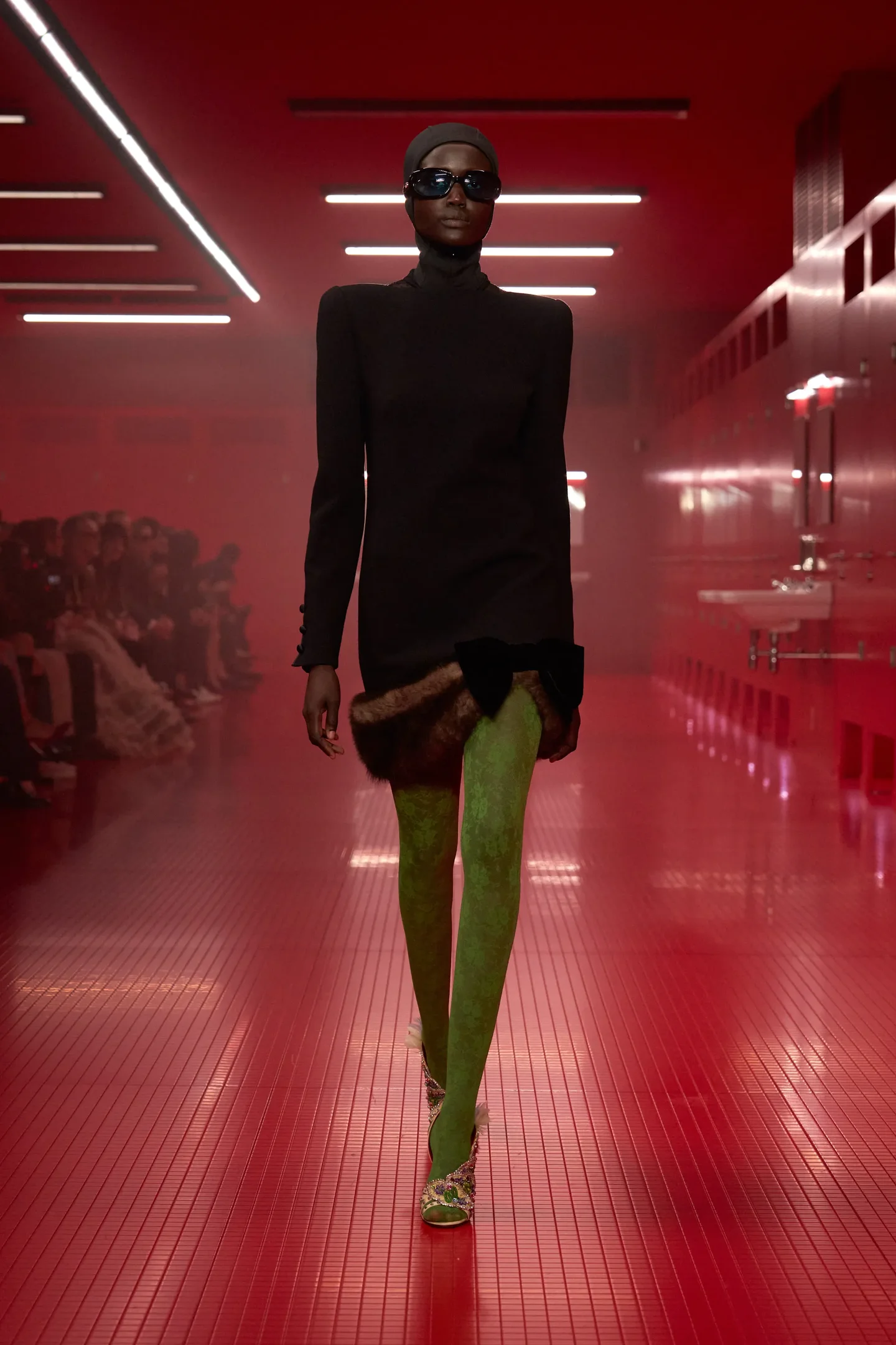 Desfile com modelo usando vestido preto e meia-calça verde em passarela vermelha, destacando estilos da temporada Outono/Inverno 25.