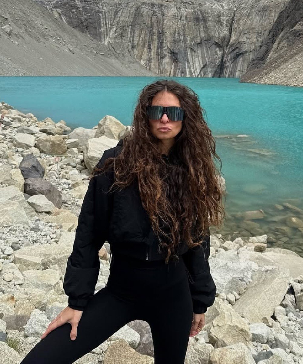 Mulher de cabelos longos e ondulados em frente a lago e montanhas, usando jaqueta e óculos escuros.