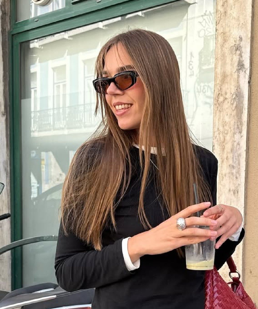 Mulher com óculos escuros e cabelo liso longo segura copo, posando à frente de vitrine.