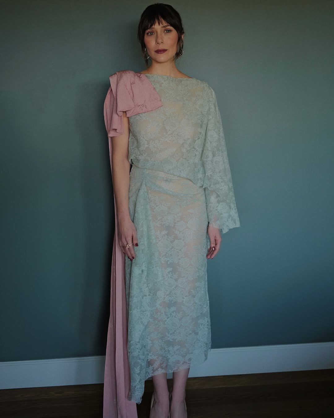 Pessoa em vestido rendado azul claro com detalhe rosa no ombro em frente a parede verde.