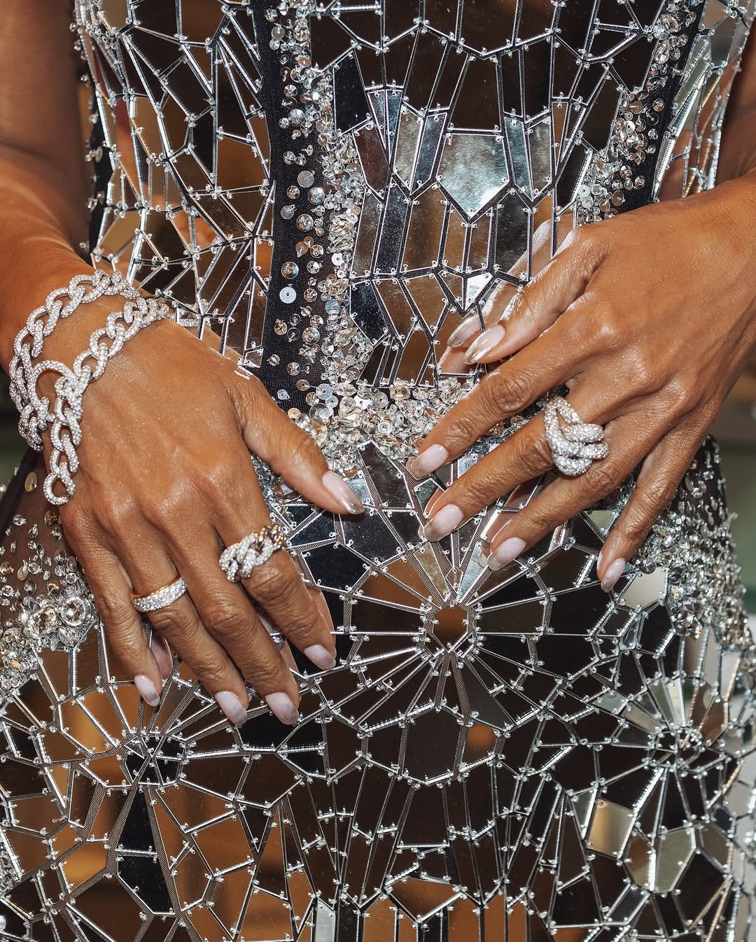 Unhas elegantes com esmalte nude, anéis e pulseira de brilho sobre vestido espelhado.