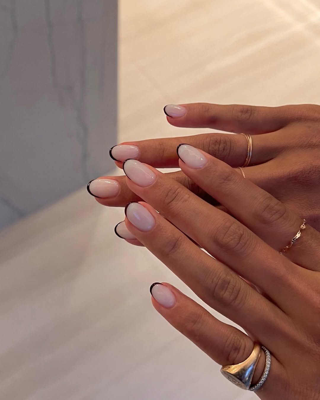 Unhas com esmalte nude e pontas francesinha preta. Mãos com anéis delicados.