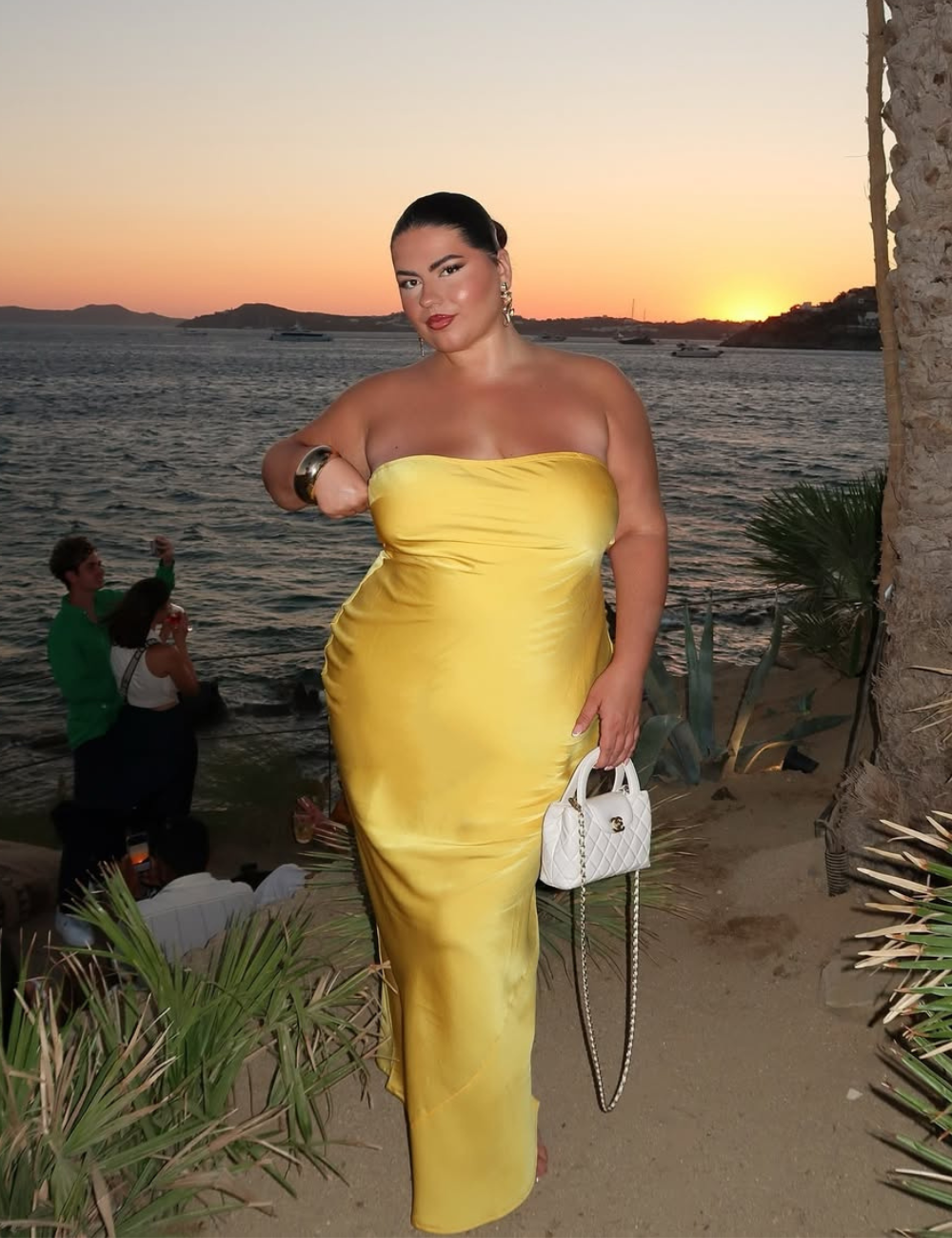 Mulher em vestido amarelo elegante à beira-mar ao pôr do sol, incorporando vestidos tendência de 2025.