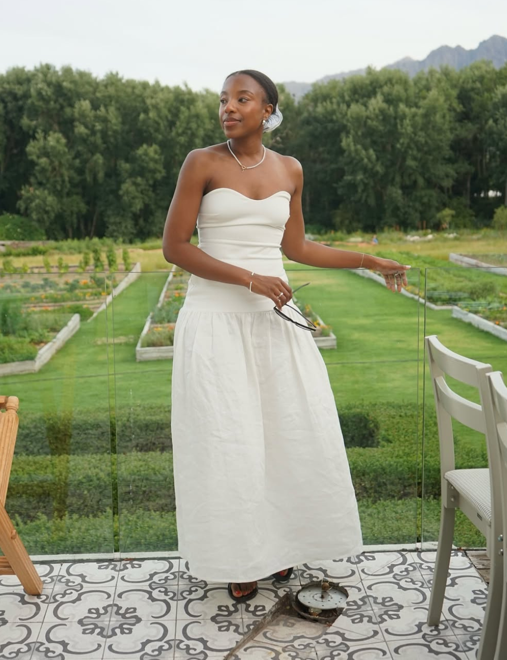 Mulher em vestido branco moderno, tendência de 2025, em varanda com jardim ao fundo.